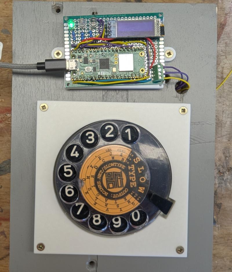 Finished Slowtype machine. A little Microconreoller with a phone dial on a grey piece of wood.